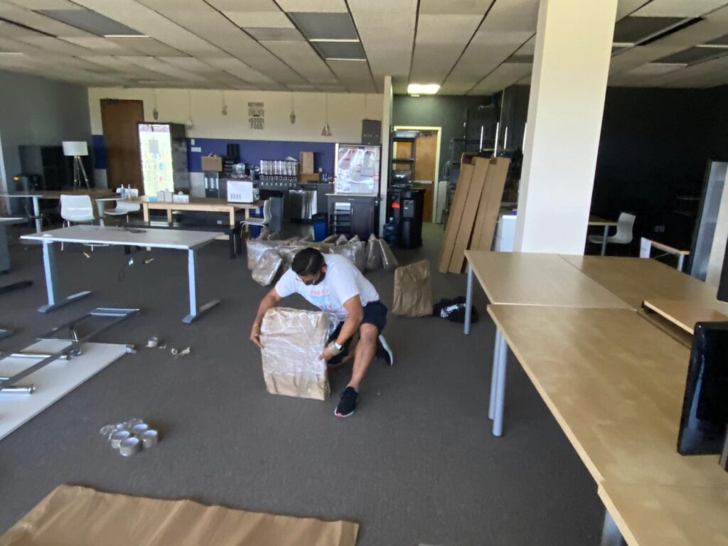 Mover wrapping items in an office with desks and packing materials around him.