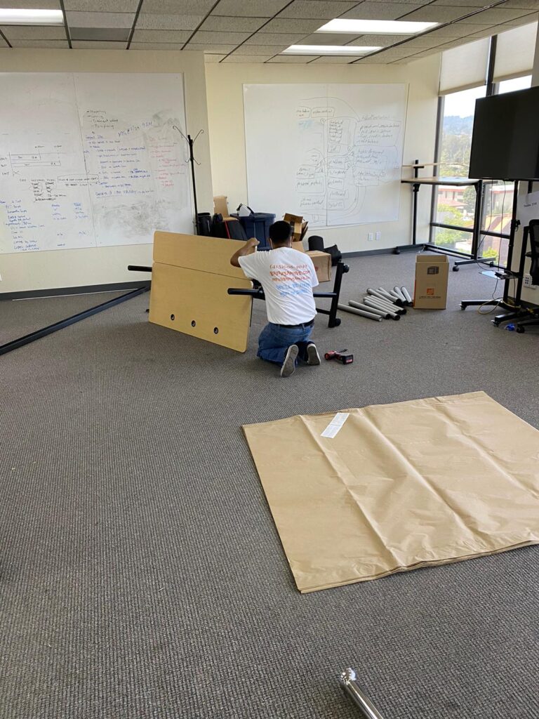 Person disassembling a table in an office space with whiteboards and a carpeted floor.